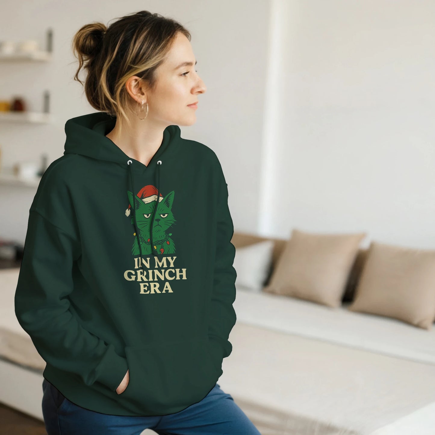 Model wearing green ‘In My Grinch Era’ hoodie with Grinch-style cat design, cozy indoor setting.
